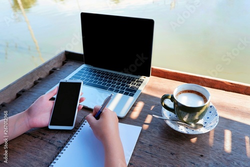 Woman hand holding smartphone and writing list of product ordered by customer with laptop and cup of coffee on wooden tabletop at waterfront house, business and technology concept photo