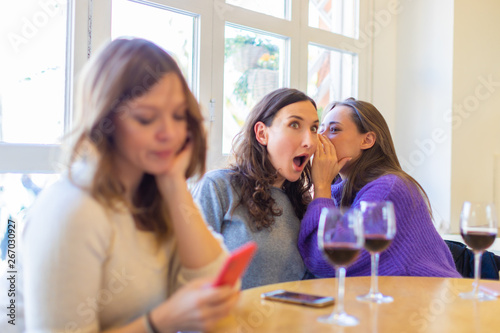Two malicious girls gossiping while their friend is disappointed looking to the phone - group of friends (women) sitting at the table with glasses of wine, female friendship and discrimination concept