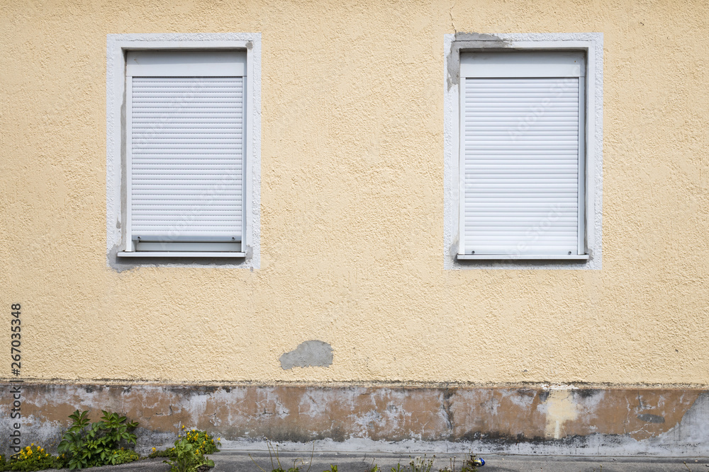 alte, runter gekommene gelbe Hauswand, mit zwei Fenstern, deren Jalousien geschlossen sind alte, runter gekommene gelbe Hauswand, mit zwei Fenstern, deren Jalousien geschlossen sind