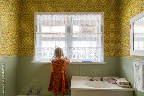 Young girl alone exploring the bathroom of a 1970s house. Lace curtains and mustard yellow colored wallpaper with green. Basin with taps and flannels. Child thinks noone is watching. Naughty.