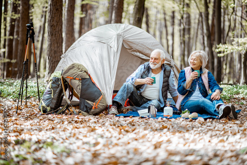 Senior couple sitting together at the campsite with tent and backpacks, enjoying nature and drinking coffee in the forest