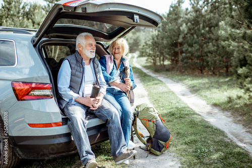 Senior couple sitting at the car trunk, enjoying nature while traveling in the young pine forest