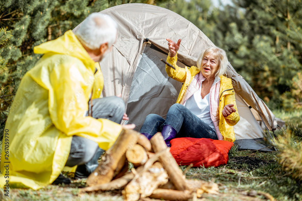 Senior couple in yellow raincoats having fun while making fireplace at the campsite near the tent in the woods Senior couple in yellow raincoats having fun while making fireplace at the campsite near the tent in the woods