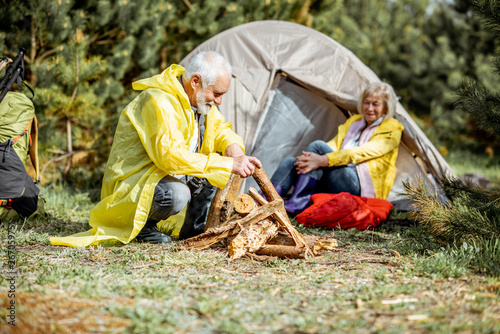 Senior couple in yellow raincoats making fireplace at the campsite near the tent in the woods