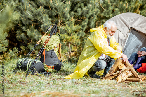 Senior man in yellow raincoat making fireplace at the campsite near the tent in the woods