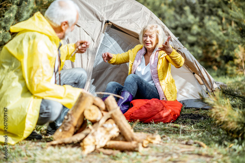 Senior couple in yellow raincoats having fun while making fireplace at the campsite near the tent in the woods