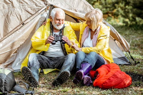 Senior couple in yellow raincoats sitting together near the tent at the campsite in the woods