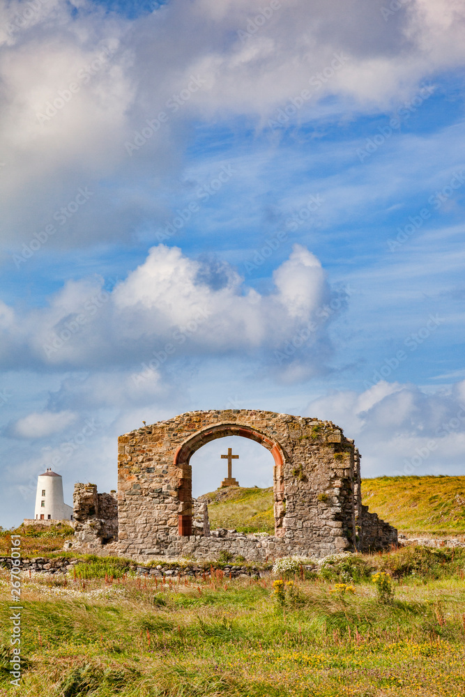 The ruined Church of St Dwynwyn, looking through a window to a large  cross, and the old lighthouse, Twr Mawr, on the tidal Island of Llanddwyn, Anglesey, Wales, UK