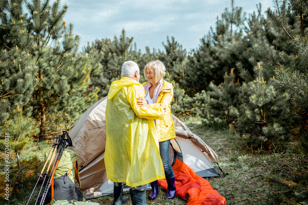 Lovely senior couple in yellow raincoats hugging together while camping with tent in the pine forest Lovely senior couple in yellow raincoats hugging together while camping with tent in the pine forest