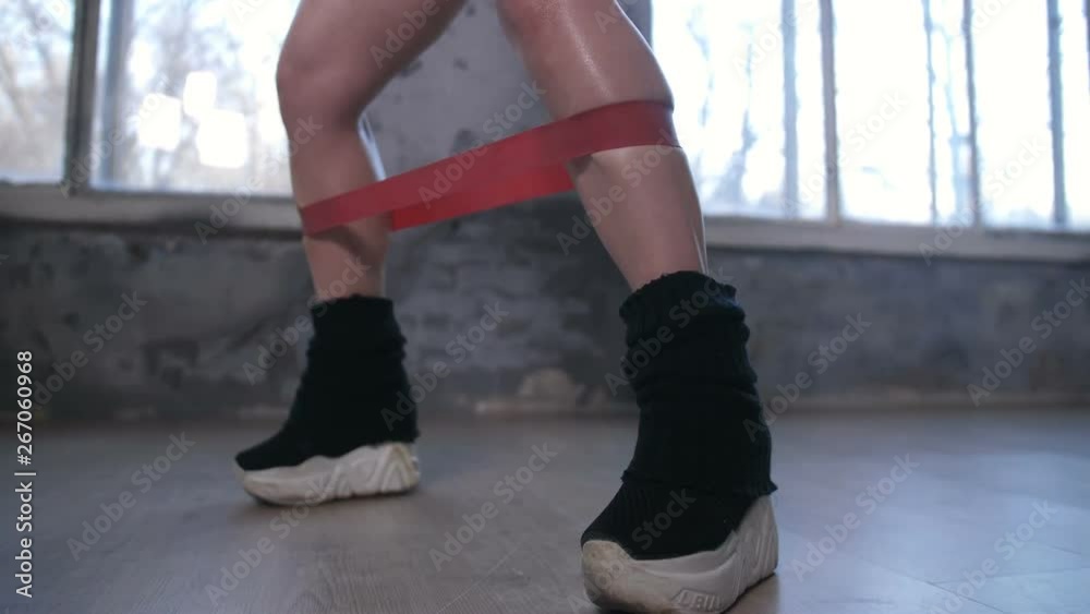 Side view of female trained legs in sports shoes and gaiters tied with resistance band during fitness workout in modern gym. Athletic woman making sport exercises with rubber band. Dolly shot Side view of female trained legs in sports shoes and gaiters tied with resistance band during fitness workout in modern gym. Athletic woman making sport exercises with rubber band. Dolly shot