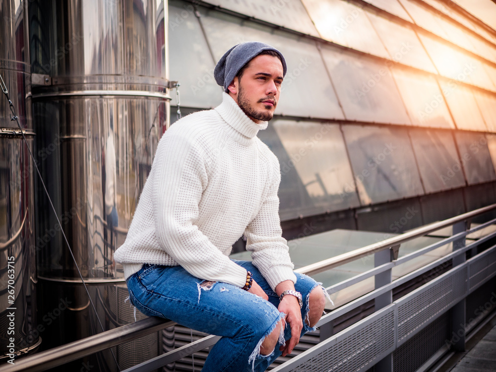 One attractive man in urban setting in modern city, sitting on metal handrail, wearing beanie cap and wool sweater, looking away