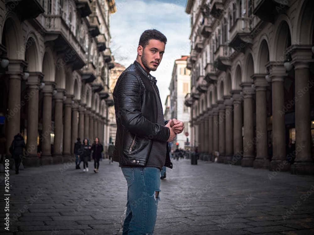 One handsome young man in urban setting in old classic European city, standing, wearing black leather jacket and jeans, looking at camera in Turin, Italy