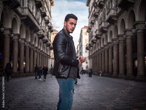 One handsome young man in urban setting in old classic European city, standing, wearing black leather jacket and jeans, looking at camera in Turin, Italy
