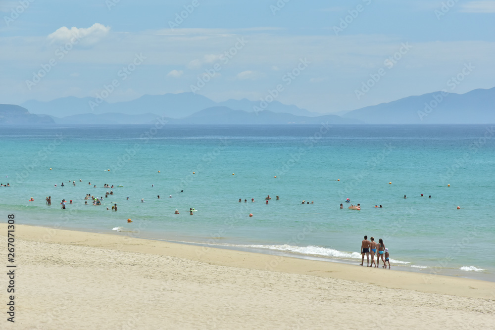 Beautiful European clean white sand beach with blue water against the background of the cloudy sky and mountains. Family of four people walking along the coast