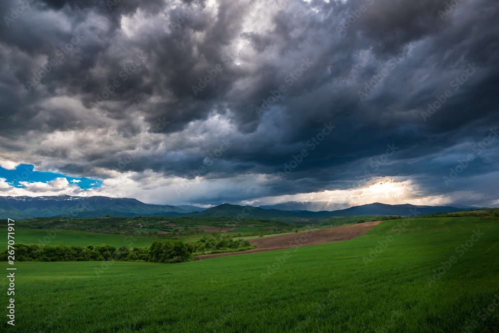 A beautiful nature scenic of a green field valley with some bush and distant mountain range under a dramatic clouds sky at sunset