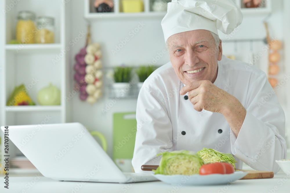 Portrait of elderly male chef cooking at table with laptop Portrait of elderly male chef cooking at table with laptop