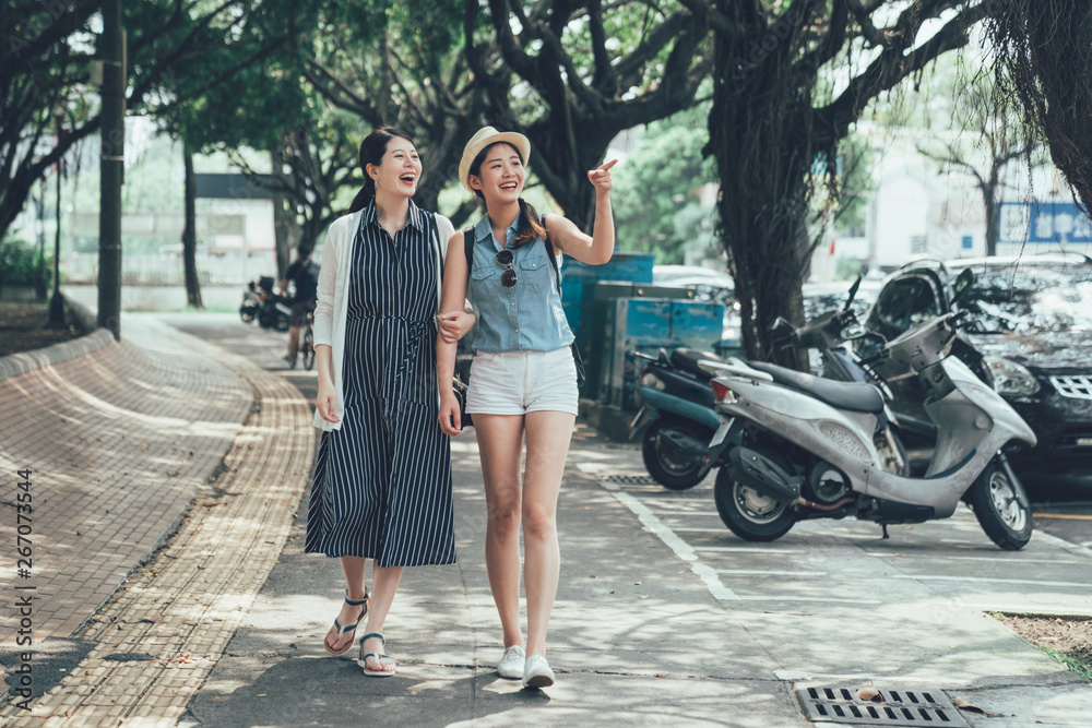 Naklejka premium two young asian girls walking on city street under trees shadow. full length beautiful female friends travelers relax outdoor excited point finger sharing amazing view in urban travel taiwan taipei