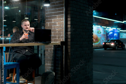 Business man with the laptop uses a mobile communication in cafe. Night time view from the street. Dark theme, in black style. Guy is chatting, working, blogging. Social media. Online education.