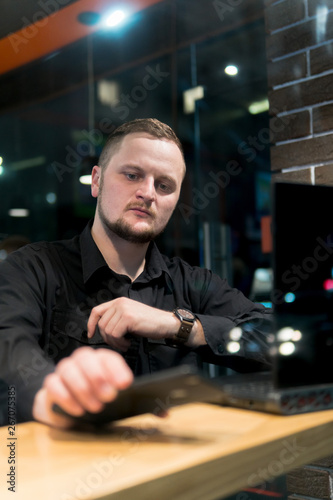 Business man looks at the phone  holds it in his hand in cafe. Night time view from the street. Dark theme  in black style. Guy is chatting  working  blogging. Social media. Online education. vertical