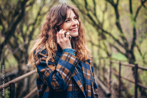 Elegant young woman with curly hair smiling and talking on smartphone. Portrait of beautiful girl outdoors. Mobile communications and lifestyle concept.