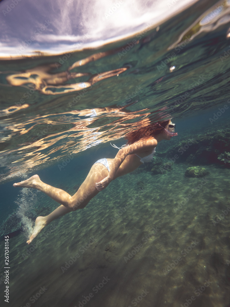 Beautiful woman is swimming diving among coral reefs in shallow water and looking underwater sealife, side view.