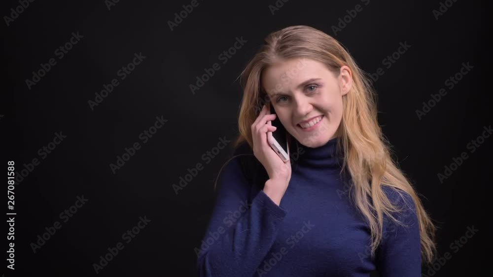Blonde model having a pleasant voicecall on smartphone watching smilingly into camera on black background. Blonde model having a pleasant voicecall on smartphone watching smilingly into camera on black background.