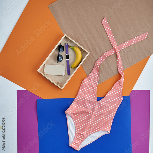 Beautiful unusual outfit presentation for fashion blog, advertising, clothing catalog. Trendy pink swimsuit with white polka dots. Wooden box with accessories: sunglasses, watches, telephone, banana.