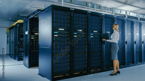 In Data Center: Female IT Technician Stands Near Rack Cabinet and Runs Maintenance Program on Laptop, Controls Operational Server's Optimal Functioning. 