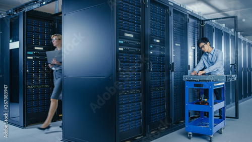 In the Modern Data Center: Team of IT Technicians Working with Server Racks, Man with Pushcart Changes Faulty Hard Drives, Doing Hardware Maintenance and Diagnostics. photo