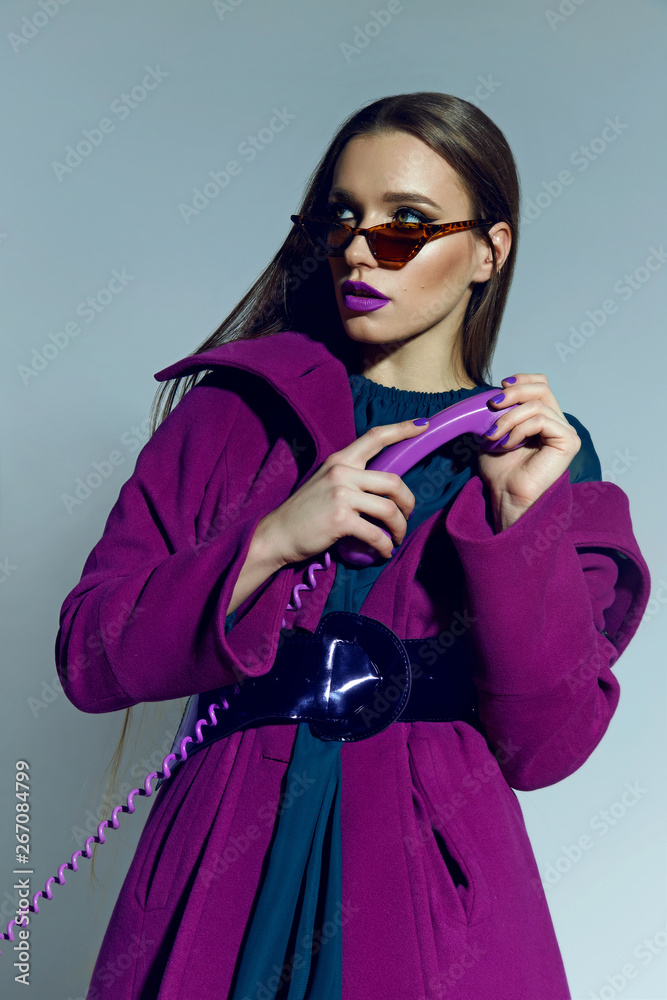 Obraz premium beautiful spectacular long-haired girl in sunglasses and a bright coat holding a lilac phone in hand. Disk phone