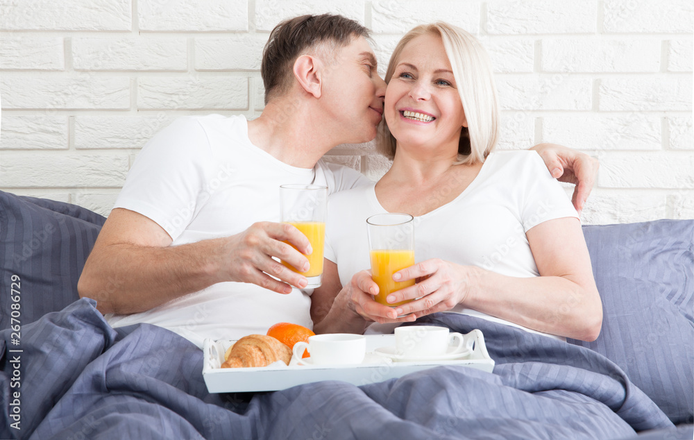 Beautiful couple having breakfast lying in bed at home. Handsome man and attractive woman are enjoying spending time together while gently hugging and kissing sitting in bed.