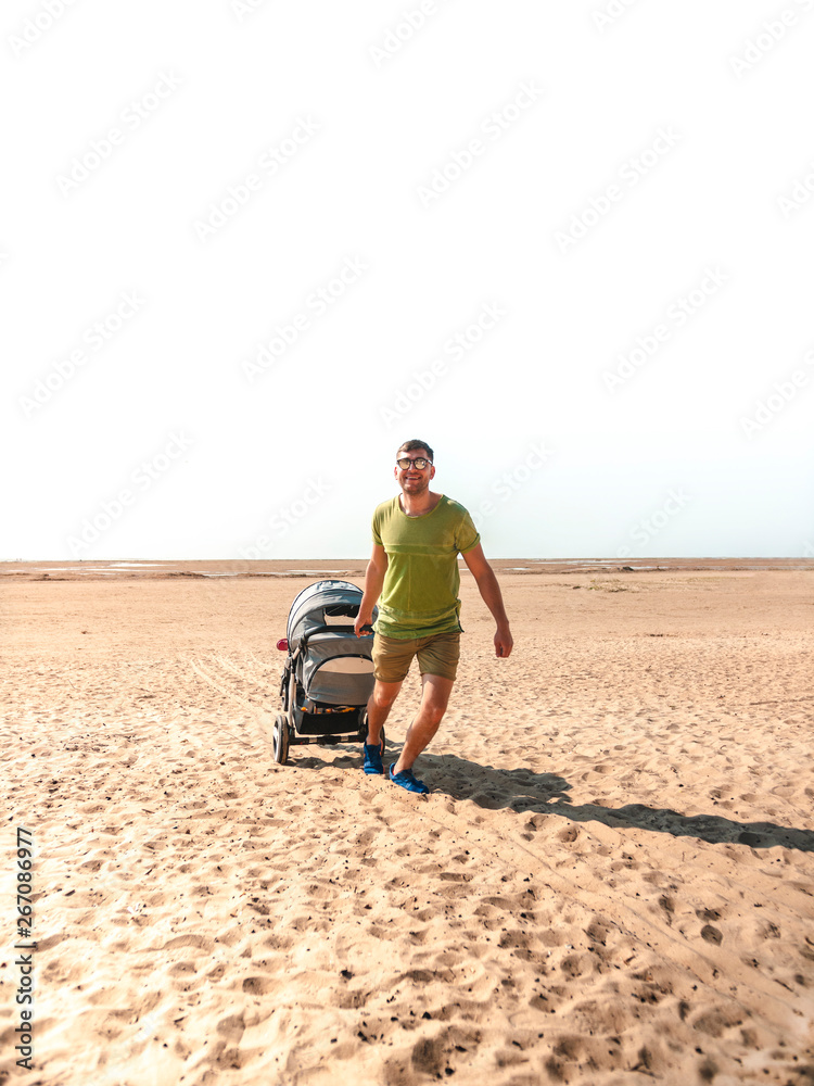 Young attractive father walks in the summer with a stroller on the sandy beach near the sea. Dad walks with his daughter. Caring young father