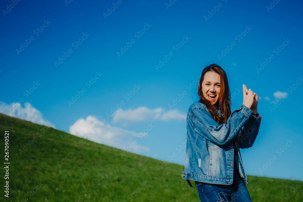 Beautiful young woman in denim jacket dancing on top of green hill on blue sky background Beautiful young woman in denim jacket dancing on top of green hill on blue sky background