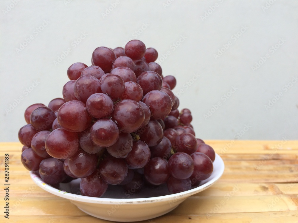 Purple grapes placed in a white plate on a bamboo table, blurred background. Purple grapes placed in a white plate on a bamboo table, blurred background.