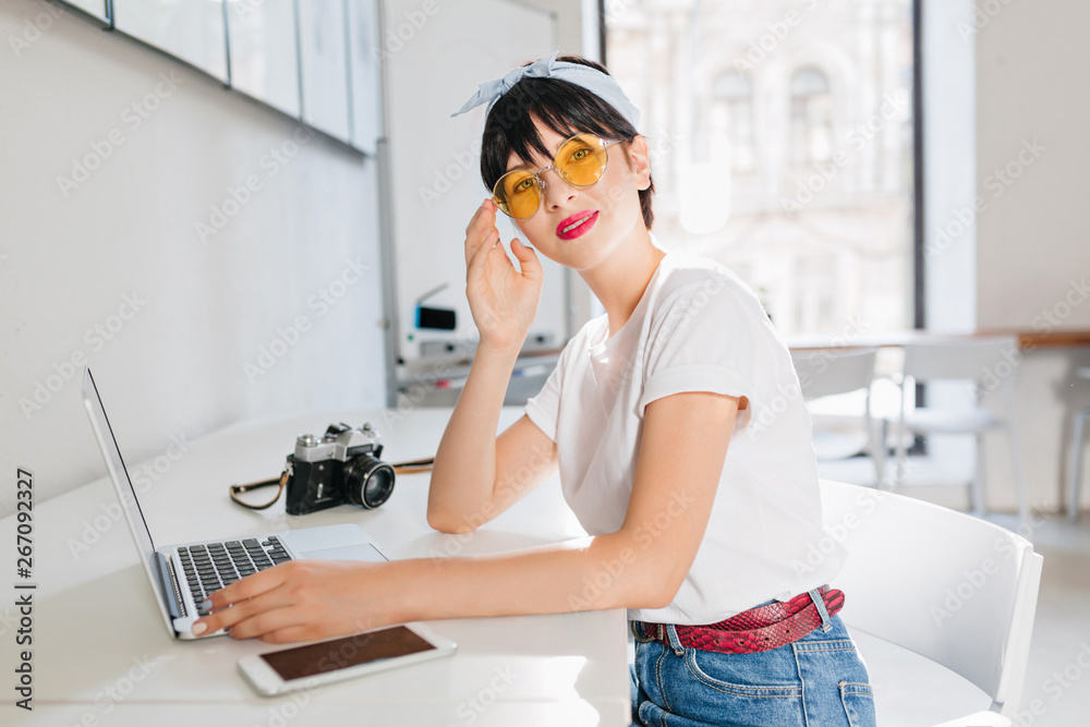 Cute brunette girl wears yellow glasses and leather belt working in office sitting with laptop and smartphone. Attractive female photographer in jeans waiting for photoshoot in studio with camera. Cute brunette girl wears yellow glasses and leather belt working in office sitting with laptop and smartphone. Attractive female photographer in jeans waiting for photoshoot in studio with camera.
