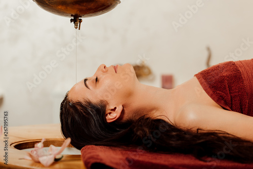 side view of relaxed young woman lying under shirodhara vessel during ayurvedic procedure photo