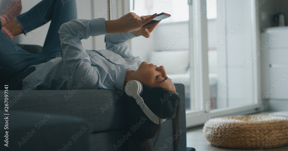 Portrait of  of an young woman is lying on the sofa is using a smart phone for listening to the music with wireless headphones in living room.