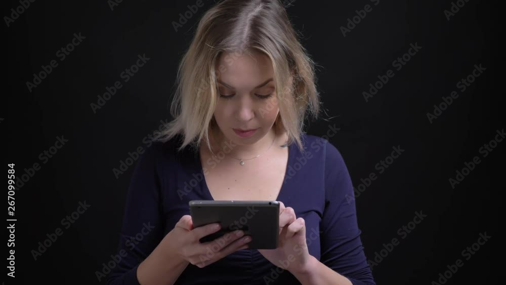 Blonde businesswoman in blue blouse watching with positive interest into tablet at black background.