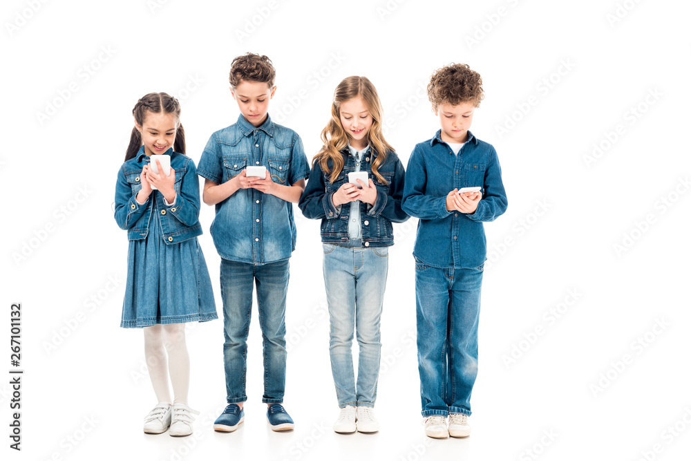 Fototapeta premium full length view of four kids in denim clothes using smartphones isolated on white