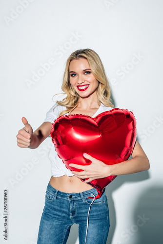 cheerful blonde girl holding red heart-shaped balloon while showing thumb up on white