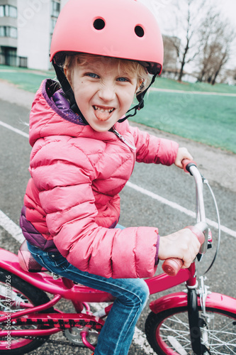 Smiling Caucasian preschooler girl riding pink bike bicycle in helmet on backyard road outside on spring day. Seasonal child activity concept. Healthy childhood lifestyle. Kid making silly faces