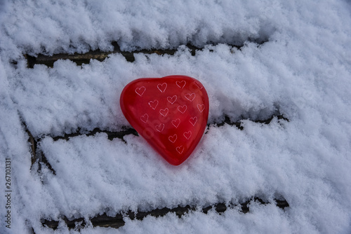 Red heart shaped stone laying on wooden table which is covered with fresh snow.