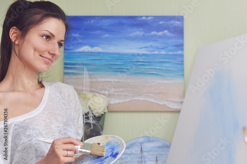A beautiful woman artist paints a picture of blue acrylic paints on a large white canvas at her home against the background of her painting of the sea.
