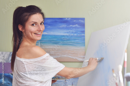 A beautiful woman artist paints a picture of blue acrylic paints on a large white canvas at her home against the background of her painting of the sea.