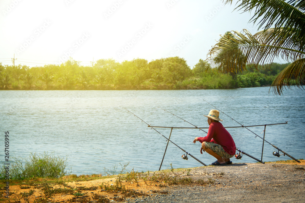 Fishing activities. fisherman middle-aged man red shirt with a straw hat  and three fishing rods on natural water swamp background landscape. Stock  Photo
