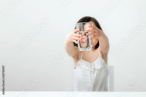 The little girl drinks water from a glass cup. The girl in a white suit drinks clear water. Girl shows us that she drinks water