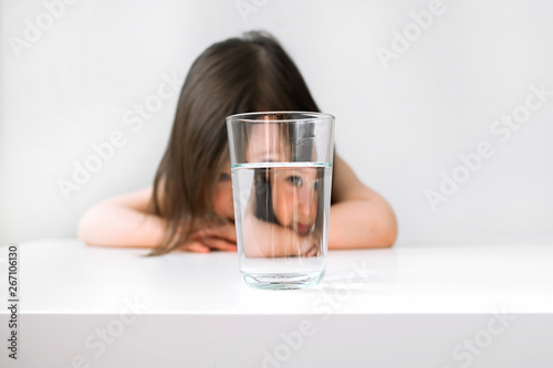 Little girl does not want to drink water. The girl sits at the table upset. The girl is sad because she does not want to drink water. Girl sitting next to a glass of water