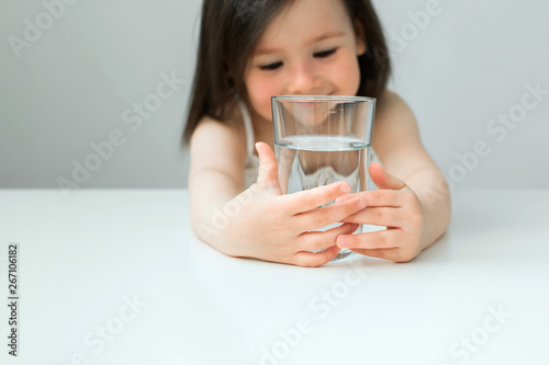 The little girl drinks water from a glass cup. The girl in a white suit drinks clear water. Girl shows us that she drinks water