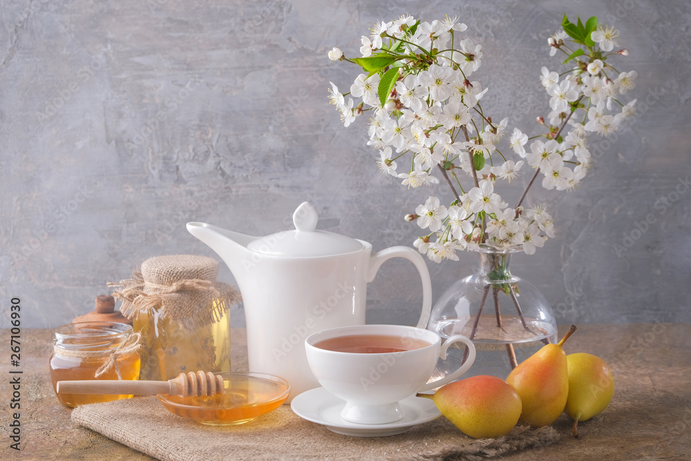A cup of tea, a white kettle, pears, jars and a saucer with honey, a bouquet of cherry blossoms on the table. Morning breakfast. A cup of tea, a white kettle, pears, jars and a saucer with honey, a bouquet of cherry blossoms on the table. Morning breakfast.