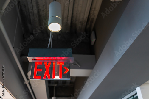 Red exit sign hanging from ceiling looking up view with arrow direction and color illuminated fixture in office building emergency closeup